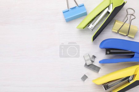 Bright staplers with staples and binder clips on light wooden table, flat lay. Space for text
