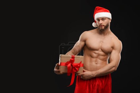 Photo for Attractive young man with muscular body holding Christmas gift box on black background, space for text - Royalty Free Image