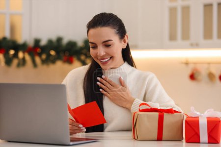 Weihnachten online feiern und Geschenke per Post austauschen. Lächelnde Frau liest Grußkarte und Geschenke während Videoanruf zu Hause