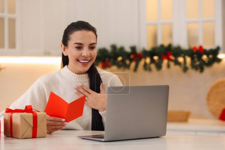 Celebrating Christmas online with exchanged by mail presents. Smiling woman with greeting card and gifts during video call at home