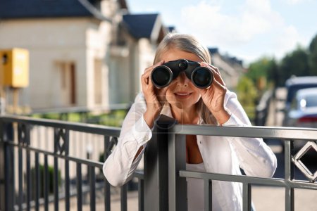 Concept de vie privée. Femme âgée curieuse avec des jumelles espionnant les voisins au-dessus de la clôture à l'extérieur