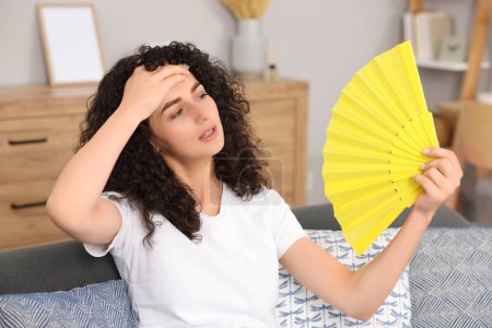 Mujer joven agitando ventilador de mano amarillo para refrescarse en el sofá en casa
