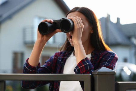 Konzept des Privatlebens. Neugierige junge Frau mit Fernglas spioniert Nachbarn über Gartenzaun aus