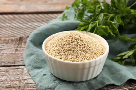 Dry quinoa seeds in bowl and parsley on wooden table, closeup