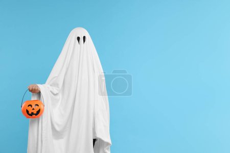Foto de Mujer en traje de fantasma blanco sosteniendo cubo de calabaza sobre fondo azul claro, espacio para el texto. Halloween celebración - Imagen libre de derechos