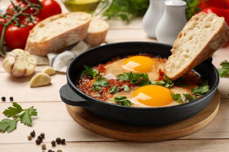 Delicious shakshuka with bread in frying pan on light wooden table, closeup