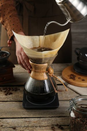 Photo for Woman pouring hot water into glass chemex coffeemaker with paper filter at wooden table, closeup - Royalty Free Image