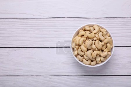 Tasty cashew nuts in bowl on white wooden table, top view. Space for text