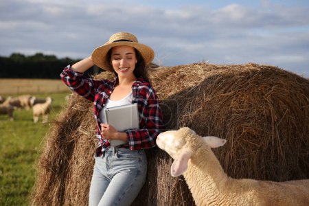 Lächelnder Bauer mit Tablet und Schafen in der Nähe von Heuballen auf Bauernhof