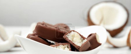 Delicious milk chocolate candy bars with coconut filling on white table, closeup. Banner design