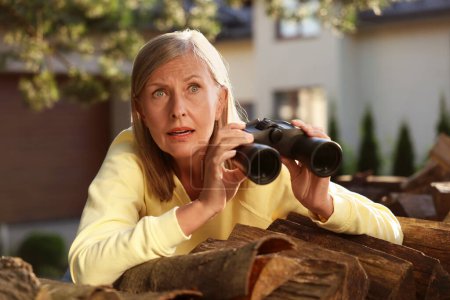 Konzept des Privatlebens. Neugierige Seniorin mit Fernglas spioniert Nachbarn über Brennholz im Freien aus