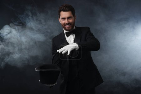 Magicien heureux montrant tour de magie avec chapeau haut de forme en fumée sur fond sombre