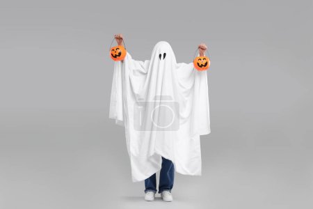 Mujer en traje de fantasma blanco sosteniendo cubos de calabaza sobre fondo gris claro. Halloween celebración