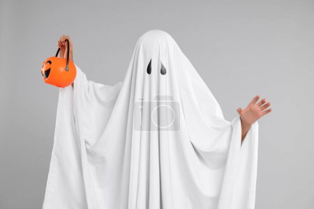 Foto de Mujer en traje de fantasma blanco sosteniendo cubo de calabaza sobre fondo gris claro. Halloween celebración - Imagen libre de derechos