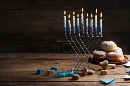 Chanukka-Feier. Menora mit brennenden Kerzen, Dreidels und Donuts auf Holztisch, Platz für Text