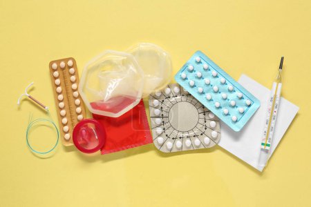 Contraceptive pills, condoms, intrauterine device and thermometer on yellow background, flat lay. Different birth control methods