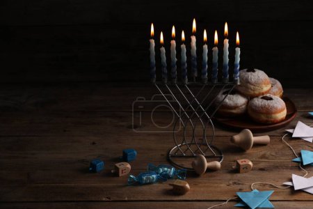 Chanukka-Feier. Menora mit brennenden Kerzen, Dreidels und Donuts auf Holztisch, Platz für Text