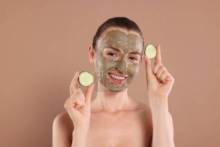 Beautiful woman with clay mask holding pieces of cucumber on light brown background