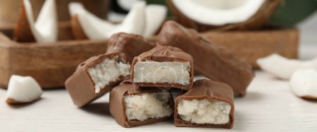 Delicious milk chocolate candy bars with coconut filling on white wooden table, closeup. Banner design