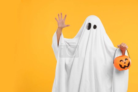 Foto de Mujer en traje de fantasma blanco sosteniendo cubo de calabaza sobre fondo amarillo, espacio para el texto. Halloween celebración - Imagen libre de derechos
