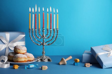 Chanukka-Feier. Menora mit brennenden Kerzen, Dreidels, Donuts und Geschenkboxen auf hellblauem Tisch, Platz für Text