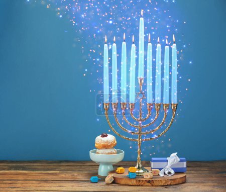 Hanukkah celebration. Menorah with burning candles, dreidels, gift box and donuts on wooden table against light blue background, space for text