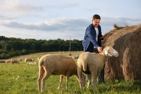 Smiling farmer with tablet stroking sheep near hay bale on animal farm. Space for text