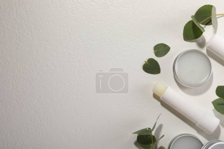 Different lip balms and leaves on white textured background, flat lay. Space for text