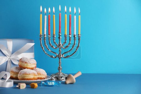 Hanukkah celebration. Menorah with burning candles, dreidels, donuts and gift box on light blue table, space for text-stock-photo