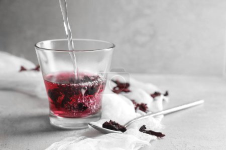 Foto de Preparando té aromático de hibisco. Verter agua en un vaso con cálices roselles secos y cuchara sobre una mesa ligera, espacio para el texto - Imagen libre de derechos