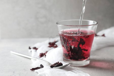 Foto de Preparando té aromático de hibisco. Verter agua en un vaso con cálices roselles secos y cuchara sobre una mesa ligera, espacio para el texto - Imagen libre de derechos