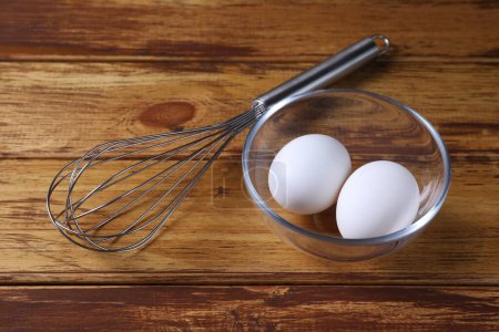 Metal whisk and eggs in bowl on wooden table, closeup