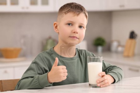 Téléchargez les photos : Garçon mignon avec verre de lait frais montrant pouce vers le haut à la table blanche dans la cuisine - en image libre de droit