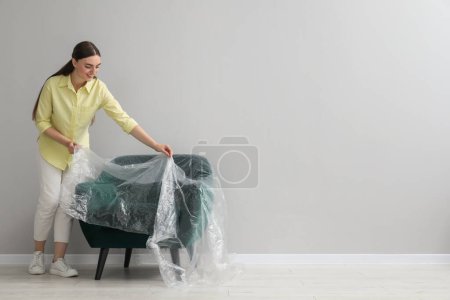 Foto de Mujer joven poniendo película de plástico lejos de sillón cerca de la pared gris claro en el interior. Espacio para texto - Imagen libre de derechos