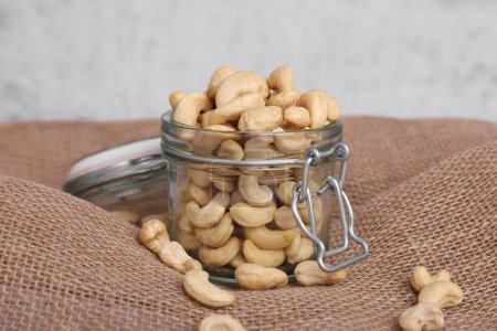 Tasty cashew nuts in glass jar on light brown fabric, closeup