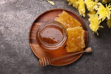 Miel d'or sucré en pot, trempette, morceaux de nid d'abeille et fleurs de chrysanthème sur table grise texturée, plat