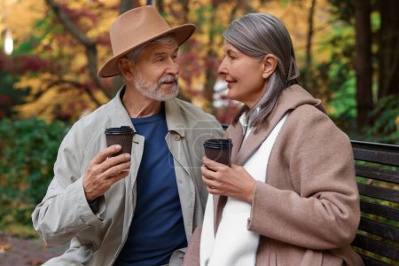 Foto de Cariñosa pareja de ancianos con tazas de café pasar tiempo juntos en el banco en el parque de otoño - Imagen libre de derechos