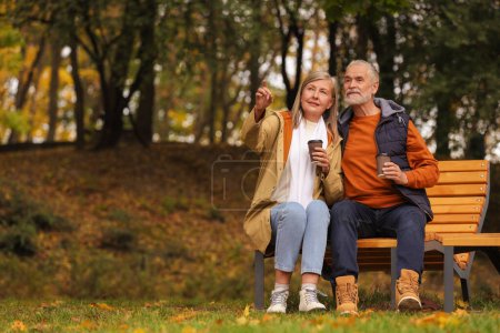 Foto de Pareja mayor cariñosa con tazas de café en el banco de madera en el parque de otoño, espacio para el texto - Imagen libre de derechos