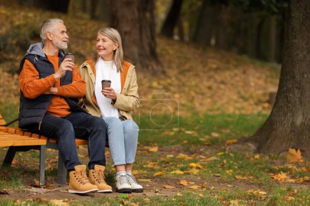 Foto de Pareja mayor cariñosa con tazas de café en el banco de madera en el parque de otoño, espacio para el texto - Imagen libre de derechos