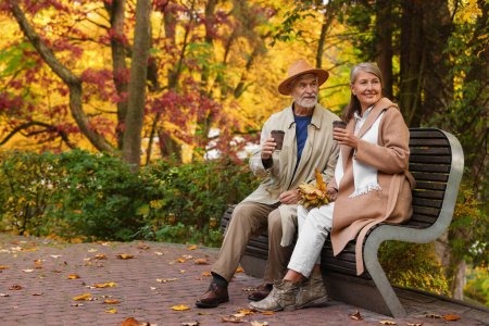 Foto de Cariñosa pareja de personas mayores con tazas de café pasar tiempo juntos en el banco en el parque de otoño, espacio para el texto - Imagen libre de derechos