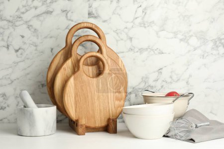 Foto de Cortadoras de madera, vajilla y utensilios de cocina sobre mesa blanca cerca de la pared de mármol - Imagen libre de derechos
