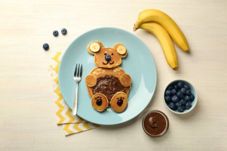 Foto de Servicio creativo para niños. Placa con oso lindo hecha de panqueques, arándanos, plátanos y pasta de chocolate en la mesa de madera ligera, puesta plana - Imagen libre de derechos