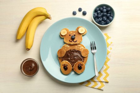 Foto de Servicio creativo para niños. Placa con oso lindo hecha de panqueques, arándanos, plátanos y pasta de chocolate en la mesa de madera ligera, puesta plana - Imagen libre de derechos