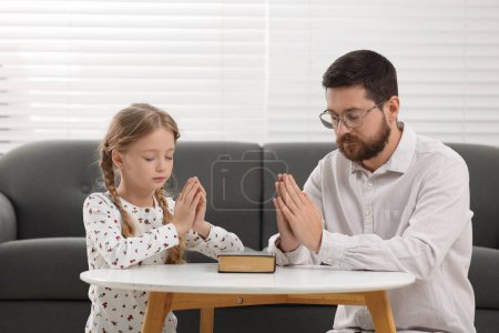 Photo for Girl and her godparent praying over Bible together at table indoors - Royalty Free Image