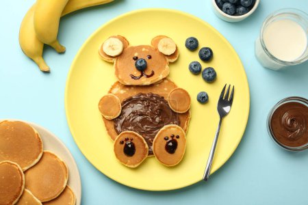 Foto de Servicio creativo para niños. Placa con oso lindo hecha de panqueques, arándanos, plátanos y pasta de chocolate en la mesa azul claro, puesta plana - Imagen libre de derechos