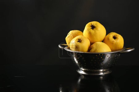 Tasty ripe quinces with water drops in metal colander on black background, space for text