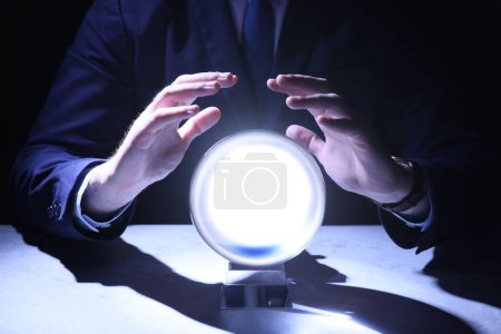 Photo for Businessman using crystal ball to predict future at table in darkness, closeup - Royalty Free Image