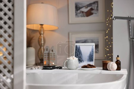 Téléchargez les photos : Plateau en bois blanc avec tablette, produits de spa et bougies allumées sur la baignoire dans la salle de bain - en image libre de droit