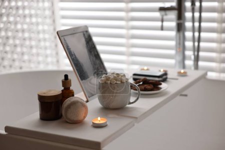 Téléchargez les photos : Plateau en bois blanc avec tablette, produits de spa, boisson chaude et bougie allumée sur la baignoire dans la salle de bain - en image libre de droit