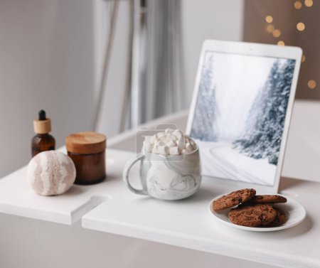 Téléchargez les photos : Plateau en bois blanc avec tablette, biscuits et produits de spa sur baignoire dans la salle de bain - en image libre de droit